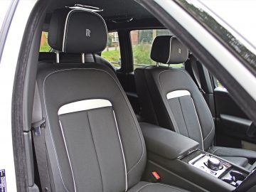 Binnenaanzicht van een Rolls-Royce Cullinan Black Badge met twee zwartleren stoelen met witte accenten, een middenconsole met bedieningselementen en het iconische Rolls-Royce-embleem op de hoofdsteunen.