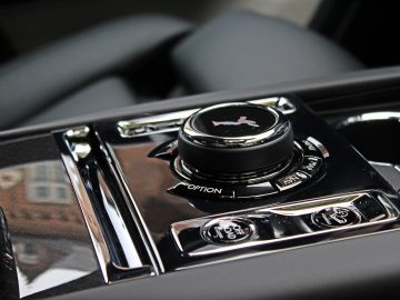 Close-up van de bedieningsknop en knoppen van een Rolls-Royce Cullinan Black Badge op een glanzend zwart paneel. De knop heeft een embleem van een staande figuur en functies voor opties, media en voertuiginstellingen.