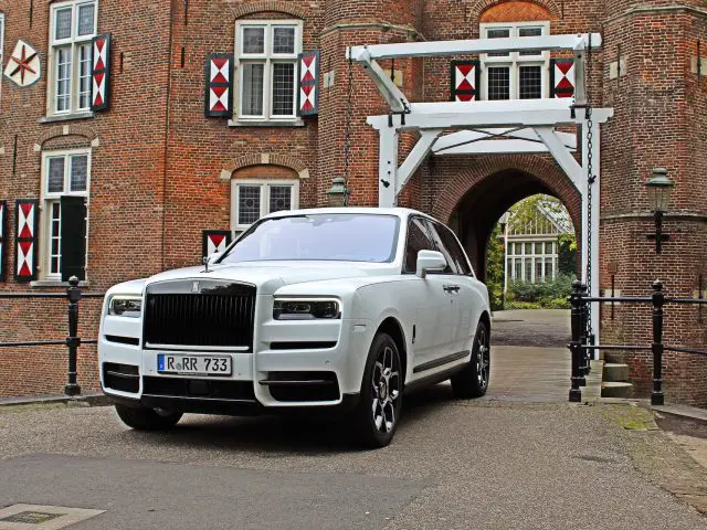 Een witte Rolls-Royce Cullinan Black Badge SUV staat geparkeerd bij een bakstenen gebouw met rode en witte luiken, voor een gebogen poort met een witte bovenconstructie.