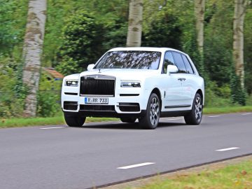 Een witte Rolls-Royce Cullinan Black Badge glijdt moeiteloos over een met bomen omzoomde weg.