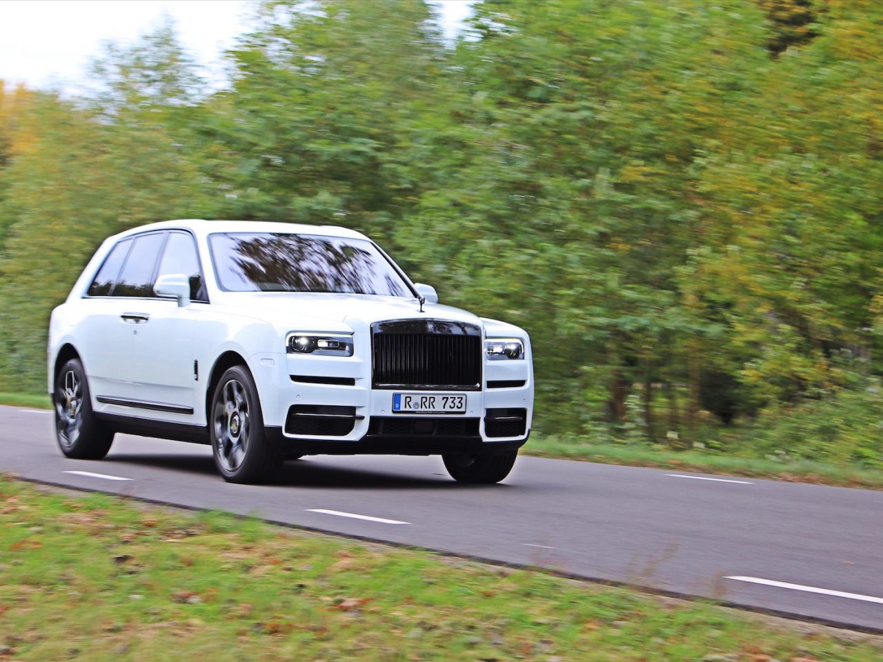 Een Rolls-Royce Cullinan Black Badge glijdt moeiteloos over een landelijke weg omgeven door weelderig groen en straalt een aura uit van ongeëvenaarde luxe en verfijning.