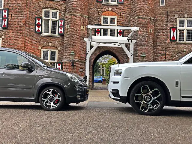Een kleine grijze auto en een grote Rolls-Royce Cullinan Black Badge staan tegenover elkaar geparkeerd in een straat, voor een historisch bakstenen gebouw met een centrale boog.
