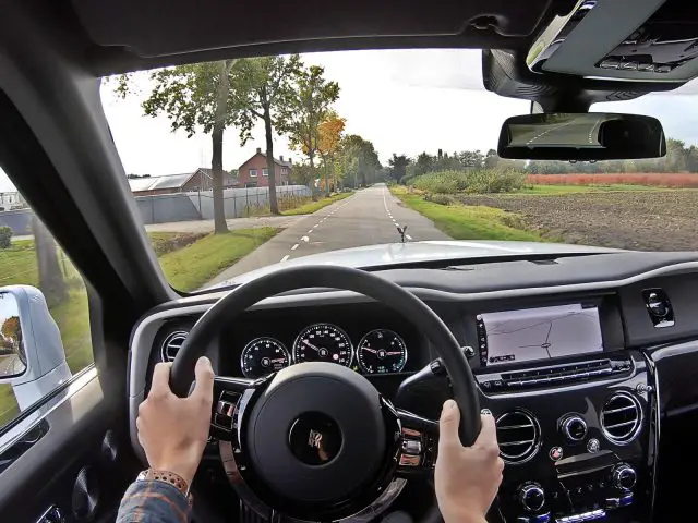 Een persoon rijdt in een Rolls-Royce Cullinan Black Badge over een landelijke weg, met de handen aan het stuur. Het dashboard en navigatiescherm zijn zichtbaar. Door de voorruit zijn velden en huizen te zien.