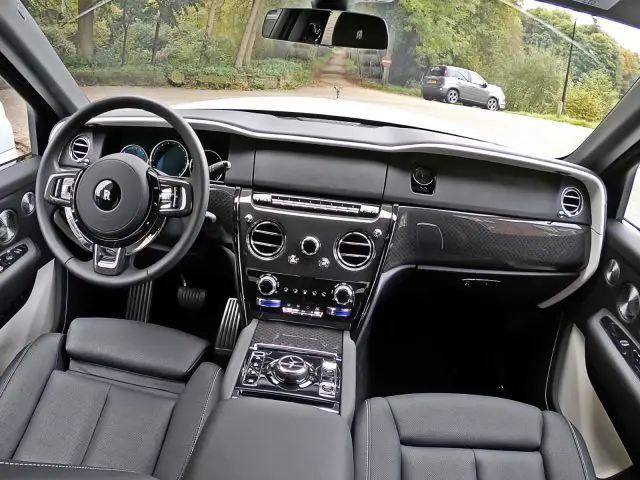 Binnenaanzicht van een Rolls-Royce Cullinan Black Badge met een strak dashboard, lederen stoelen, multifunctioneel stuur en een middenconsole met verschillende bedieningselementen. Bomen en andere geparkeerde auto's zijn door de ramen zichtbaar.