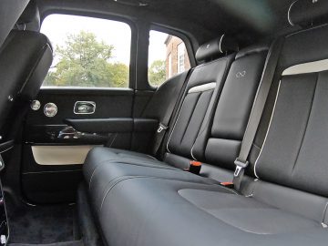 De afbeelding toont het interieur van een Rolls-Royce Cullinan Black Badge, met een ruime achterbank met zwart lederen bekleding, witte accenten en voldoende beenruimte. Bovendien beschikt de auto over grote ramen en een indrukwekkend zicht naar buiten.