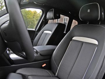 Interieur van een Rolls-Royce Cullinan Black Badge met zwartleren stoelen met witte accenten, zichtbare stoelmarkering op de hoofdsteunen en een deel van het dashboard van het voertuig. Buiten zijn bomen en een gebouw zichtbaar.