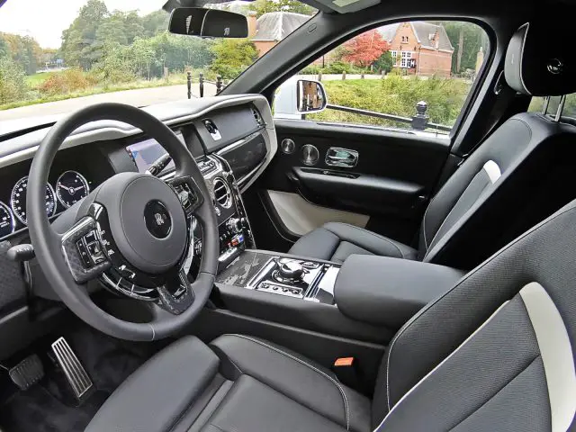 Interieur van de Rolls-Royce Cullinan Black Badge met een leren stuur, digitaal dashboard, centrale bedieningsconsole en comfortabele zwarte stoelen met witte accenten. Door de ramen is een schilderachtig uitzicht zichtbaar.