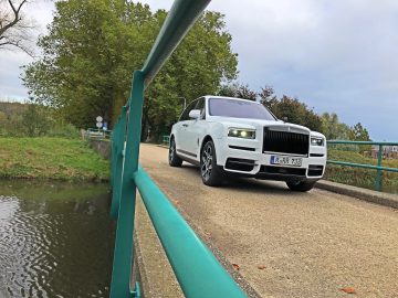 Een Rolls-Royce Cullinan Black Badge met Europese kentekenplaten rijdt op een smalle brug over een kleine watermassa, groen en bomen op de achtergrond.