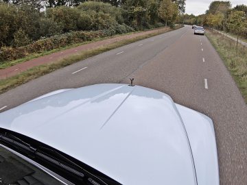 Een lichtgekleurde Rolls-Royce Cullinan Black Badge rijdt op een tweebaansweg met aan de linkerkant een fietspad en aan beide kanten grasvelden. Andere voertuigen zijn verderop op dezelfde weg zichtbaar.