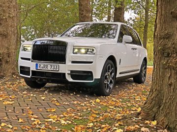 Een witte Rolls-Royce Cullinan Black Badge luxe SUV met een Europees kenteken staat geparkeerd op een geplaveid pad omgeven door bomen met gevallen bladeren.