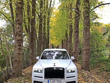 Een witte Rolls-Royce Cullinan Black Badge luxe SUV staat geparkeerd op een geplaveid pad omzoomd met hoge bomen met vergeelde bladeren in een serene bosrijke omgeving.