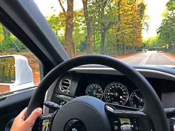 Uitzicht vanaf de bestuurdersstoel van een Rolls-Royce Cullinan Black Badge die op een zonnige dag over een met bomen omzoomde weg rijdt, met het luxueuze stuur, het dashboard en de snelheidsmeter.