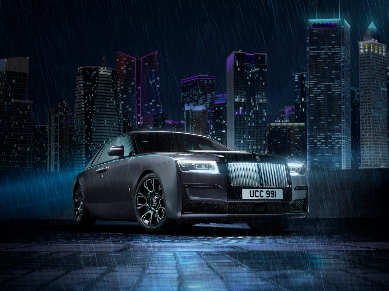 Een luxe Rolls-Royce Ghost Black Badge met het kenteken "UCC 991" staat geparkeerd op een nat oppervlak in een stadsgezicht tijdens zware regenval 's nachts, met verlichte wolkenkrabbers op de achtergrond.