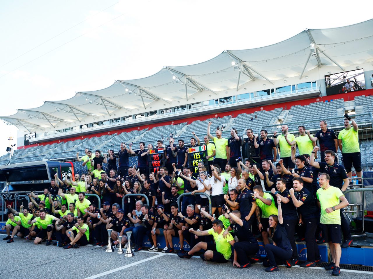 Een grote groep mensen poseerde samen op een racebaan, sommigen hielden trofeeën vast en anderen staken hun armen omhoog. De achtergrond bestaat uit lege toeschouwerstribunes en een overdekte zithoek, waardoor de opwinding van de overwinningsviering van Max Verstappen wordt vastgelegd.