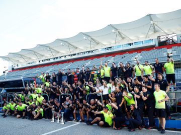 Een grote groep mensen poseerde samen op een racebaan, sommigen hielden trofeeën vast en anderen staken hun armen omhoog. De achtergrond bestaat uit lege toeschouwerstribunes en een overdekte zithoek, waardoor de opwinding van de overwinningsviering van Max Verstappen wordt vastgelegd.