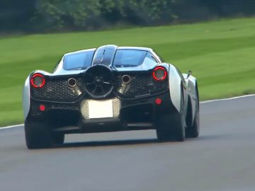 Een T.50 sportwagen met opvallende achterlichten en aerodynamisch design rijdt op een weg met aan beide kanten groen gras.