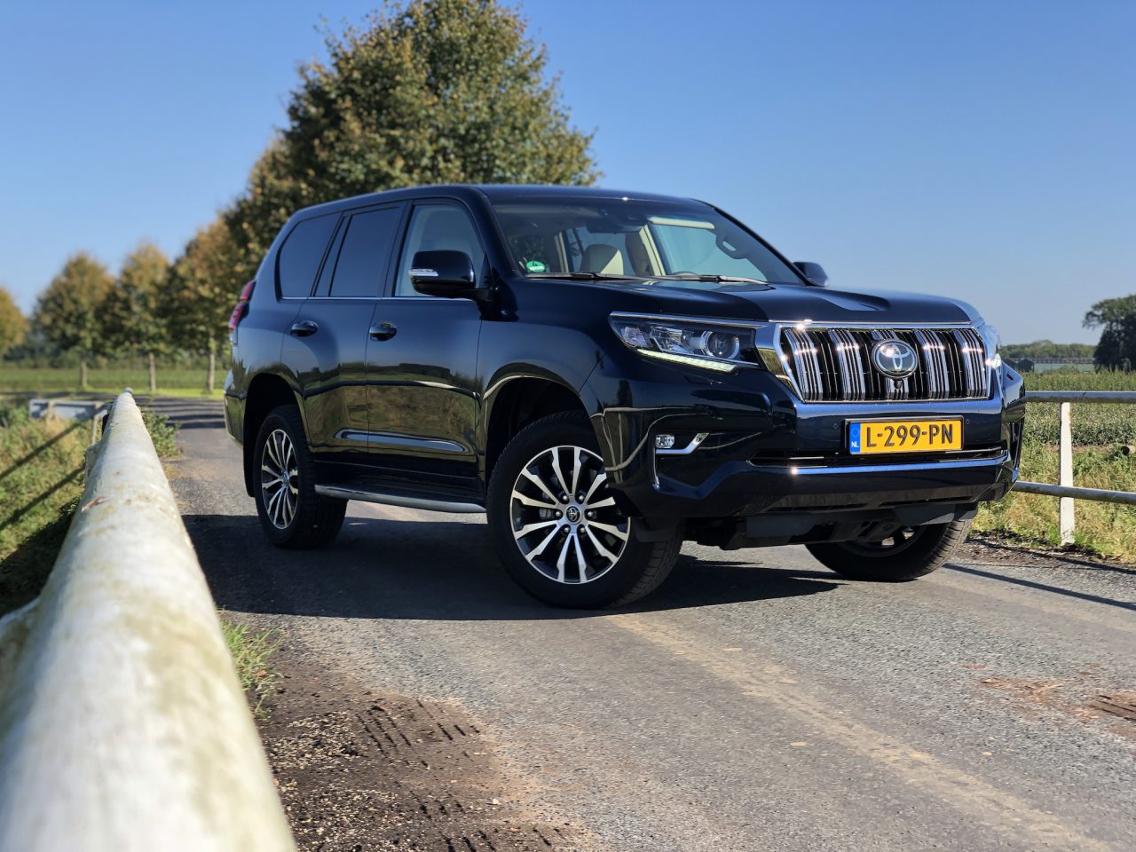 Een zwarte Landcruiser met Nederlands kenteken staat geparkeerd op een landelijke weg, omgeven door bomen en weelderig groen op de achtergrond.