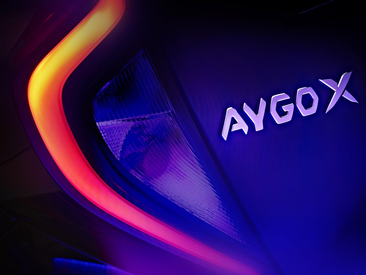 Close-up van het achterlicht en "AYGO X"-badge van een Toyota Aygo X, verlicht door rode en paarse verlichting.