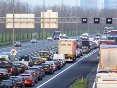 Zware verkeersopstoppingen op een snelweg met meerdere rijstroken met verschillende voertuigen, waaronder auto's en vrachtwagens, die zich als bestanden verplaatsen. Verkeersborden erboven geven snelheidslimieten en wegmarkeringen weer. Op de achtergrond staan bomen langs de weg.