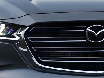 Close-up van de grille van een strakke grijze Mazda-auto, met de kenmerkende koplampen en het prominent weergegeven Mazda-embleem in het midden.