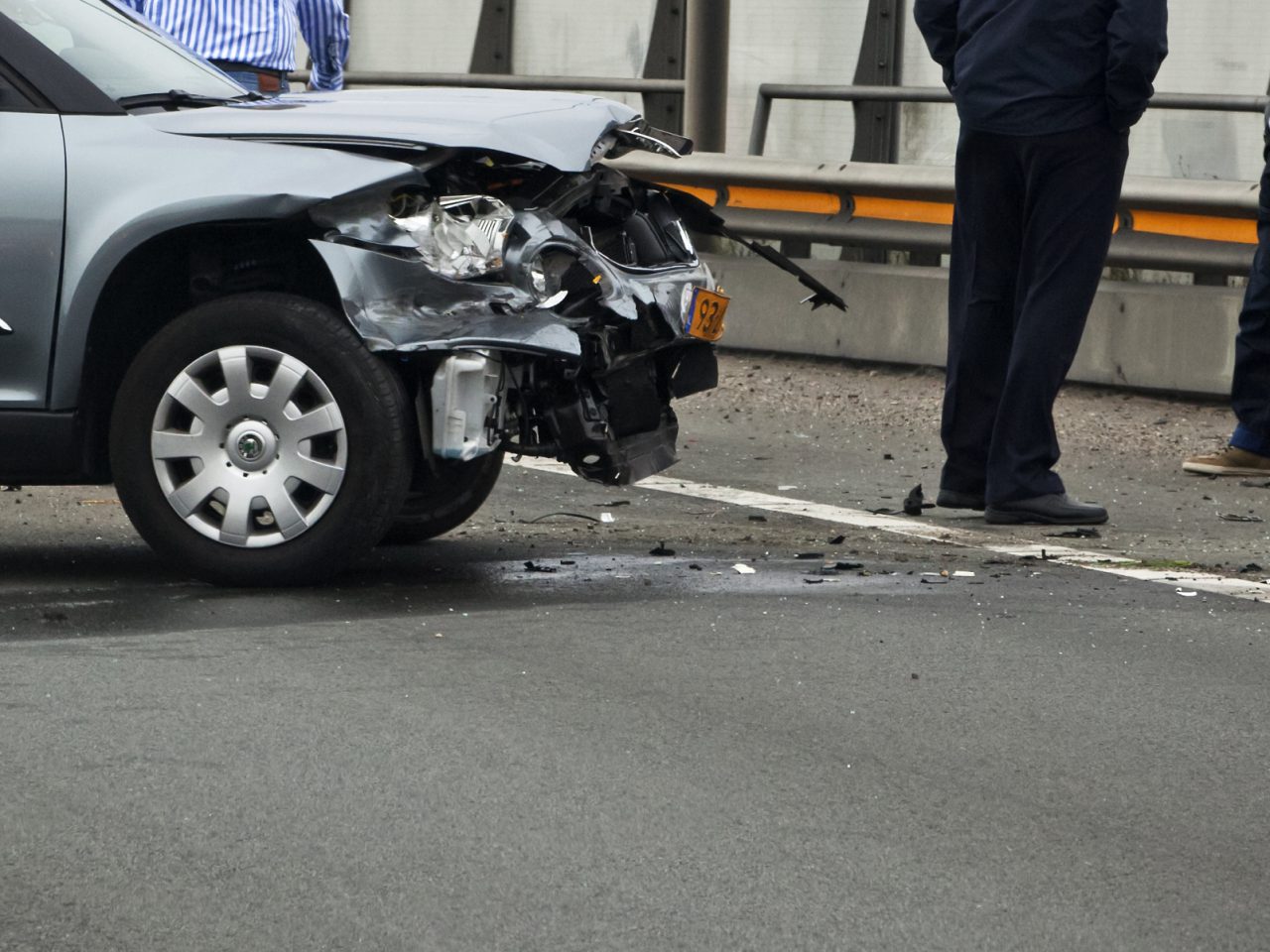 Een beschadigde auto met een verfrommelde voorbumper en motorkap staat op de weg vlakbij een slagboom, mogelijk als gevolg van verkeersongevallen. Er staan twee mensen vlakbij.