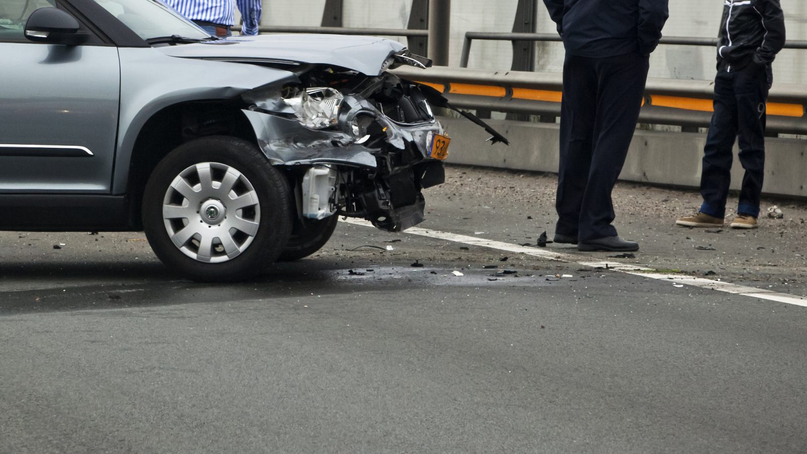 Een beschadigde auto met een verfrommelde voorbumper en motorkap staat op de weg vlakbij een slagboom, mogelijk als gevolg van verkeersongevallen. Er staan twee mensen vlakbij.