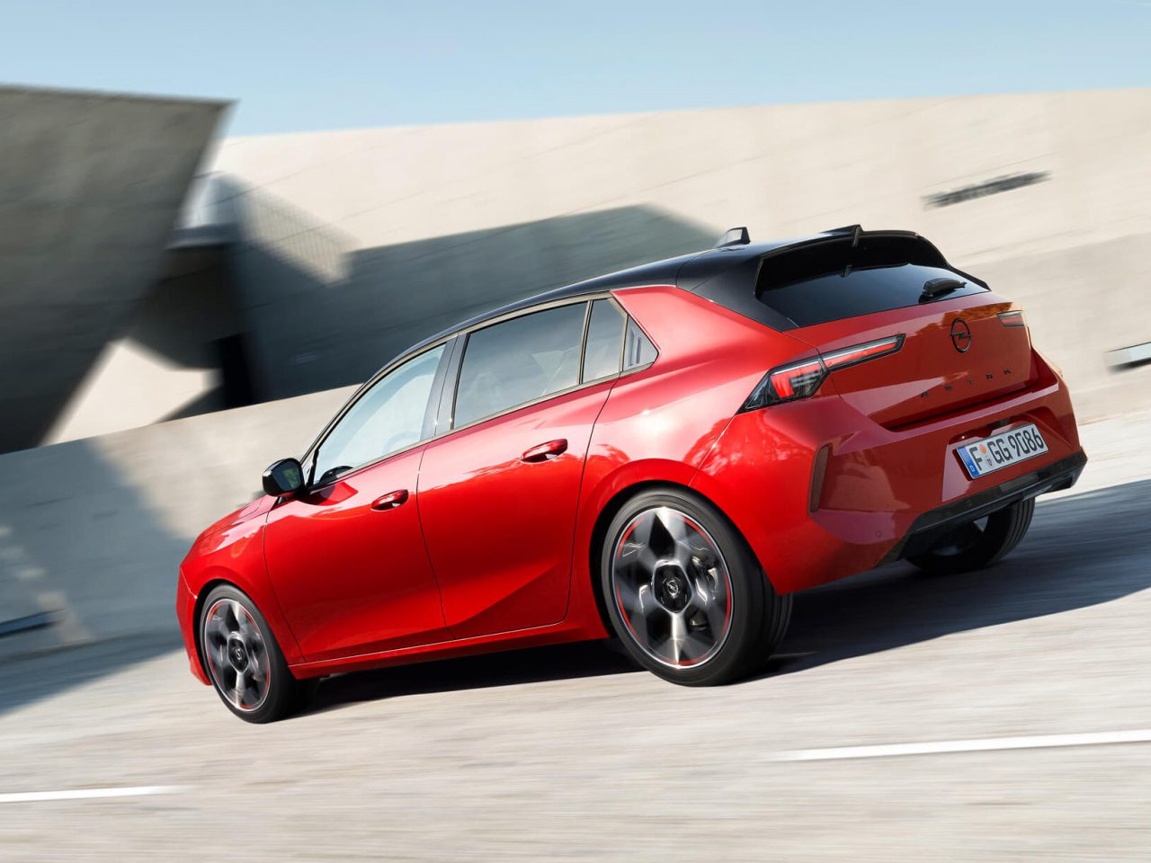 Een rode, moderne Opel Astra hatchback rijdt op een weg vlakbij een betonconstructie.