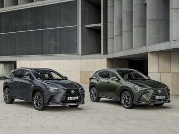 Twee Lexus SUV's, waaronder een strakke Lexus NX, staan geparkeerd op een verhard terrein voor een modern gebouw met een beige en grijze buitenkant. De linker SUV is donkergrijs en de rechter is donkergroen.
