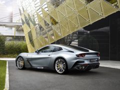 Een Ferrari BR20-sportwagen, glinsterend in zilver, staat geparkeerd op een verhard oppervlak voor een modern gebouw met gouden, ruitvormige architectuur.