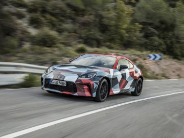 Een Toyota GR86, voorzien van een veelkleurige camouflageverf, glijdt moeiteloos over een bochtige weg omgeven door groen. Op het kenteken van de auto staat K GR 8064, wat duidelijk opvalt doordat de nieuwe Toyota trots zijn aanwezigheid kenbaar maakt als opvolger van de GT86.