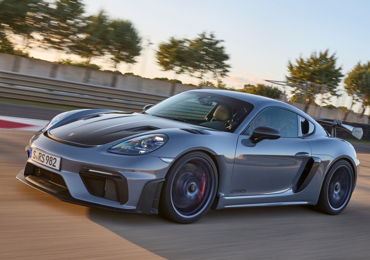 Een slanke, zilveren Porsche Cayman GT4 RS-sportwagen met een zwarte spoiler die op een racecircuit rijdt met bomen en blauwe lucht op de achtergrond.