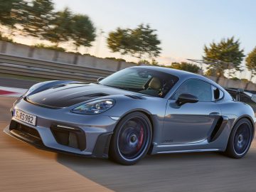 Een slanke, zilveren Porsche Cayman GT4 RS-sportwagen met een zwarte spoiler die op een racecircuit rijdt met bomen en blauwe lucht op de achtergrond.