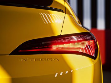 Close-up van de linkerachterkant van een gele Acura Integra met de naam "Integra" zichtbaar, met een verlicht achterlicht tegen een onscherpe achtergrond met verticale zwart-witte strepen.