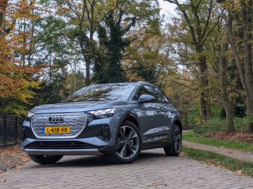 Een grijze Audi Q4 e-tron SUV met kenteken L-418-VK staat geparkeerd op een verhard pad omgeven door herfstbomen.