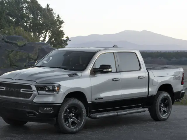 Een zilveren RAM 1500 pick-up staat geparkeerd op een verhard terrein met een rotsachtig landschap en bergen op de achtergrond.