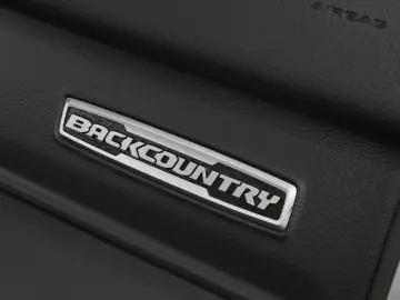 Close-up van een voertuiginterieur met een 'Backcountry'-badge op een zwart gestructureerd oppervlak. Het woord "AIRBAG" is boven het embleem in reliëf aangebracht en geeft de ruige elegantie van de RAM 1500 weer.