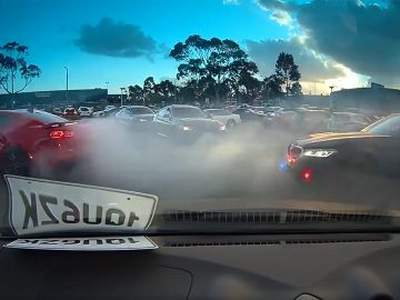 Auto's die ronddrijven en dikke rook veroorzaken op een drukke parkeerplaats tijdens zonsondergang, met duidelijk zicht op een burn-out en de kentekenplaat zichtbaar op het dashboard vanuit de auto.