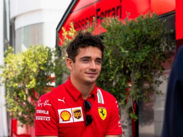 Buiten staat een persoon in een rood Ferrari-teamshirt, voor het groen en een rood gebouw met het Ferrari-logo, kijkend naar een ander individu, wat doet denken aan Charles Leclerc die zijn team observeert.