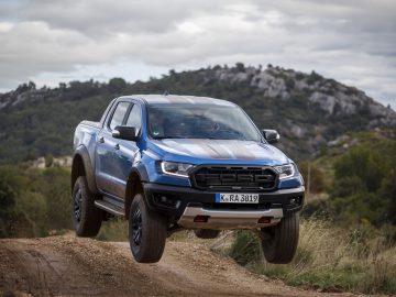Een blauwe Ford Ranger-pick-up met racestrepen vliegt in de lucht op een onverharde weg, tegen een achtergrond van heuvels en bewolkte lucht.