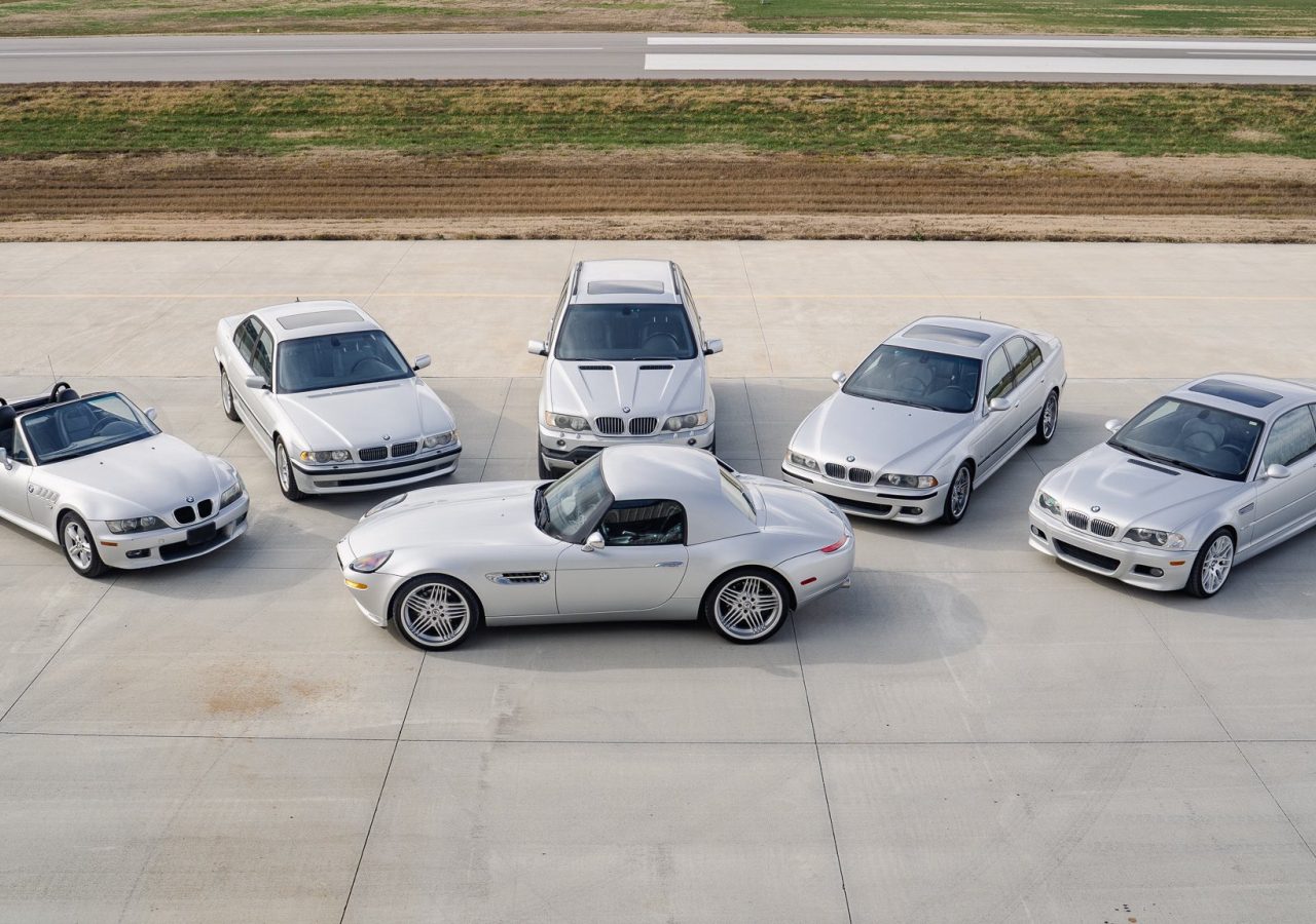 Een groep van zes strakke zilveren BMW-auto's, waaronder elegante cabriolets en luxe sedans, gerangschikt in een halve cirkel op een betonnen oppervlak met een weelderig grasveld op de achtergrond.