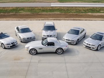 Een groep van zes strakke zilveren BMW-auto's, waaronder elegante cabriolets en luxe sedans, gerangschikt in een halve cirkel op een betonnen oppervlak met een weelderig grasveld op de achtergrond.