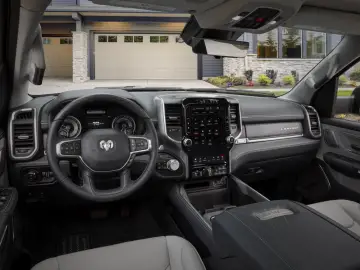 Binnenaanzicht van een moderne RAM 1500 met het stuur, het digitale instrumentenpaneel en een groot centraal touchscreen-display in het dashboard, met aan de buitenkant een woongarage en oprit zichtbaar.