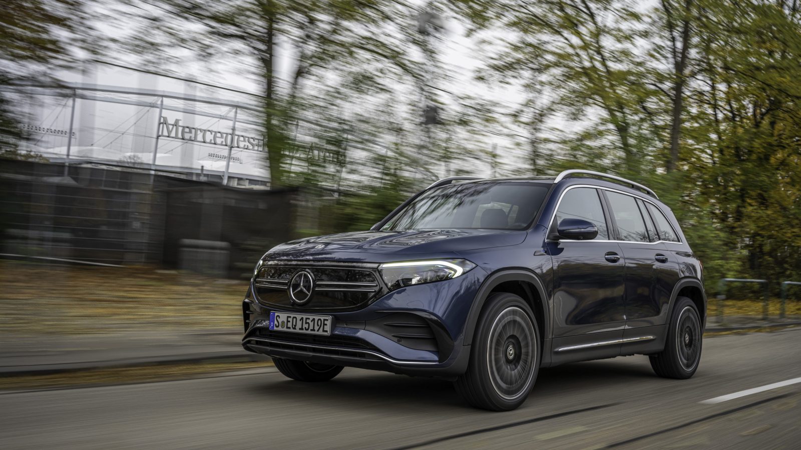 Een donkerblauwe Mercedes-Benz EQB SUV rijdt op een weg met bomen en een hek op de achtergrond.