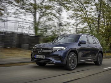 Een donkerblauwe Mercedes-Benz EQB SUV rijdt op een weg met bomen en een hek op de achtergrond.