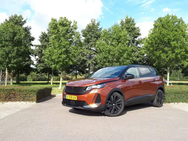 Een bruine Peugeot 5008 SUV staat geparkeerd op een parkeerplaats in de buurt van een park met bomen op de achtergrond onder een blauwe lucht met verspreide wolken. Het voertuig heeft zwarte wielen en een geel kenteken.