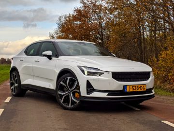 Een strak witte Polestar 2 met Nederlands kenteken "J-538-DG" staat geparkeerd op een weg met herfstbomen op de achtergrond.