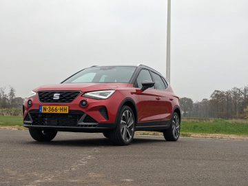 Een rode SEAT Arona-auto met Nederlands kenteken N-366-HH staat geparkeerd op een verhard oppervlak, tegen een achtergrond van een bewolkte lucht en bomen. Als u overweegt een Seat Arona-recensie te lezen, legt deze scène het stijlvolle ontwerp en de praktische aantrekkingskracht vast.