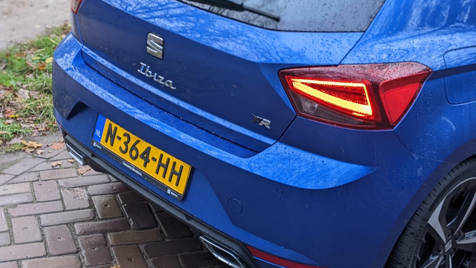 Close-up van de achterkant van een blauwe SEAT Ibiza FR met Nederlands kenteken N-364-HH, geparkeerd op een natte bakstenen stoep.
