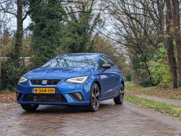 Op een kasseienpad in een bosrijke omgeving, omgeven door bomen en struiken, staat een blauwe Seat Ibiza met kenteken N-364-HH geparkeerd.