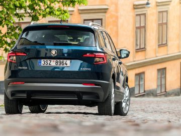 In een charmante geplaveide straat staat een blauwe Škoda Karoq geparkeerd, aangevuld met de rustieke uitstraling van een geel gebouw op de achtergrond.